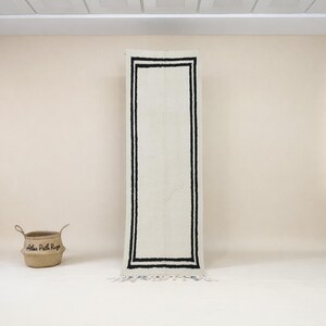 May include: A rectangular, off-white rug with a black border and fringe. The rug is displayed vertically against a light beige wall. A woven basket with the text "Atlas Path Rugs" is on the left.