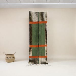 May include: A long, rectangular rug with a central green panel, bordered by black and white vertical stripes. Two horizontal orange bands cross the green section. A woven basket with handles sits to the left. The rug has a fringed edge.