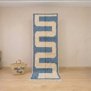 May include: A rectangular runner rug featuring a thick, blue, geometric zigzag pattern set against a cream-coloured background. The rug has a high-pile texture and is finished with short, cream-coloured fringe tassels along the top and bottom edges.