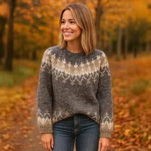 May include: A grey, beige, and white patterned jumper with long sleeves. The jumper has a round neckline and is worn with blue jeans. The background shows a blurred autumn scene with trees and fallen leaves.