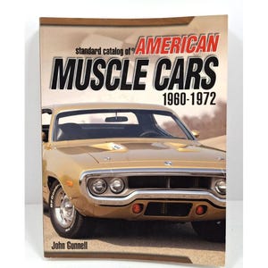 Op de afbeelding: Een boek getiteld "standard catalog of AMERICAN MUSCLE CARS 1960-1972" toont een goudkleurige klassieke auto op de cover. De auteur van het boek is John Gunnell. De titel staat in vette zwarte en rode letters.