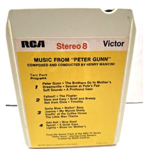 May include: Vintage RCA Victor Stereo 8-track tape of "Music From Peter Gunn" by Henry Mancini. The tape has a yellow and white label with song titles listed. The tape is in a cream-colored plastic cartridge.