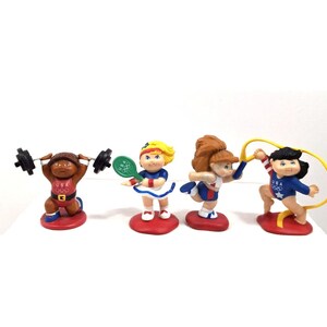 Peut inclure: Quatre figurines d'athlètes en uniforme de l'équipe olympique américaine. Les figurines sont sur des socles rouges. L'une fait de l'haltérophilie, une autre joue au tennis, une autre fait de la gymnastique et une autre utilise une corde à sauter.