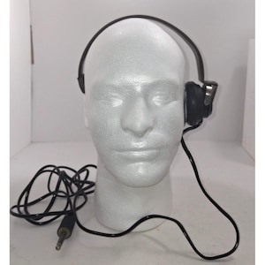 Puede incluir: Auriculares vintage negros con una almohadilla acolchada y un cable negro. Los auriculares están sobre una cabeza de espuma blanca. El cable tiene un conector jack de 0,635 cm.