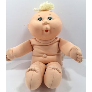 May include: A vintage Cabbage Patch Kid doll with a soft, flesh-toned body and a round face. The doll has blue eyes, rosy cheeks, and a small red mouth. A tuft of blonde yarn sits atop its head. The doll is in a seated position with arms outstretched.