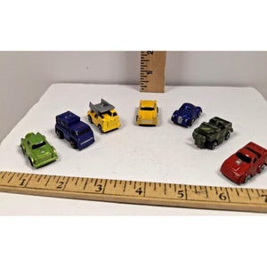 May include: A collection of eight miniature toy cars in various colors, including green, blue, yellow, and red. The cars are arranged on a white surface with a ruler in the foreground, suggesting a small scale. The toy cars are a mix of different vehicle types.