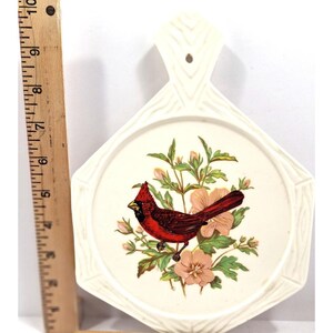 May include: Cream-coloured decorative wall hanging featuring a red cardinal bird and pink flowers. The bird is perched on a branch with green leaves. The hanging has a handle and a decorative border.