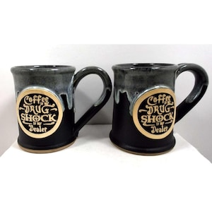 May include: Two ceramic coffee mugs with a black base and a gray top. Each mug has a large handle and a gold-colored circular emblem with the words "Coffee is my Drug Shock is my Dealer" in a stylized font.