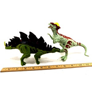 May include: Two plastic dinosaur toys. One is a green Stegosaurus with dark brown plates. The other is a green and red Dilophosaurus. A wooden ruler is placed next to the toys.