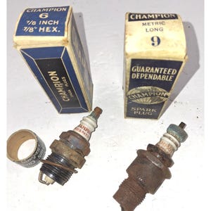 Può includere: Due candele Champion con le loro scatole originali. Una scatola recita "Champion 6 7/8 inch 7/8" HEX." L'altra scatola recita "Champion Metric Long 9." Le candele sono metalliche con isolatori bianchi e verdi.