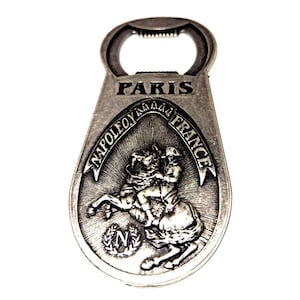 May include: Silver-toned bottle opener with a loop for leverage. The opener features the word "PARIS" at the top and an oval design with a rider on a horse, the words "NAPOLEON À FRANCE" and a laurel wreath.