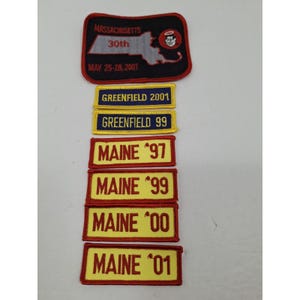 May include: A selection of embroidered patches. The top patch is black with the text "MASSACHUSETTS 30th MAY 25-28, 2001" and a state outline. Below are patches with "GREENFIELD" and "MAINE" with years.