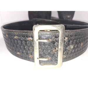 May include: Close-up of a black leather belt with a woven pattern and a silver-colored rectangular buckle. The belt shows signs of wear and tear, with some discoloration.