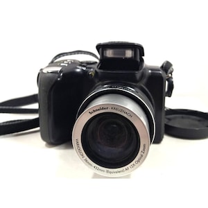 Op de afbeelding: Zwarte digitale camera met een zilveren lensring. De lens is gemarkeerd met Schneider-Kreuznach, Variogon 36mm-432mm (Equivalent) AF 12X Optical Zoom. De camera heeft een ingebouwde flitser en een zwarte riem.
