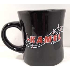 May include: Black ceramic mug with a curved handle. The mug features the text "KAMEL Cigarettes" in red letters with musical notes and lines. The mug has a glossy finish and a slightly flared shape.