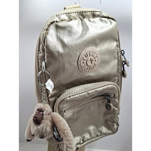 May include: A metallic gold-colored sling backpack with a small front pocket and a zipper closure. The backpack has a round logo patch and a small monkey keychain attached. The bag is made of a shiny, durable material.