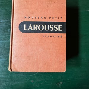 May include: A vintage book with a textured, peach-colored cover. The title "NOUVEAU PETIT LAROUSSE ILLUSTRE" is printed in black and white across the front. The book is rectangular and appears well-worn.
