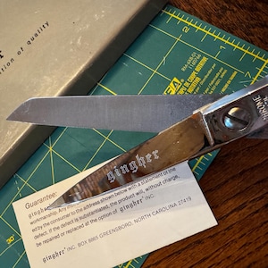 May include: A pair of chrome-plated Gingher scissors with a silver blade, resting on a green cutting mat. The scissors are next to a cardboard box with the text "gingher a tradition of quality". A guarantee is also visible.