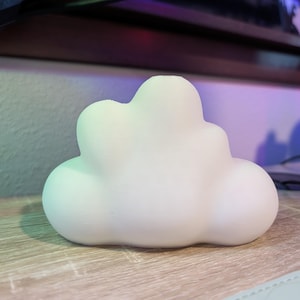 Può includere: Un oggetto decorativo bianco a forma di nuvola è appoggiato su una superficie di legno. La nuvola ha una forma liscia e arrotondata e sembra essere fatta di ceramica o un materiale simile. Lo sfondo è sfocato.