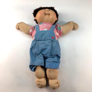 Puede incluir: Una muñeca Cabbage Patch Kid vintage con cabello castaño y mejillas rosadas. La muñeca viste una camisa de cuadros rosa y blanco con un estampado floral y un peto vaquero azul. Los brazos y las piernas de la muñeca están extendidos.