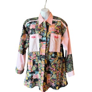 May include: A long-sleeve button-down shirt featuring a black base with a colourful farm-themed print of sunflowers, vegetables, and animals. The shirt includes solid light pink panels on the collar, cuffs, and two front chest pockets with buttons.