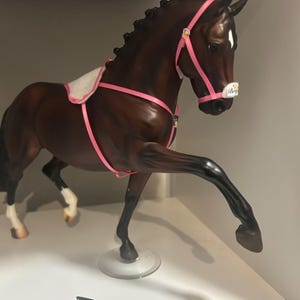 Puede incluir: Figura de caballo marrón con brida y silla de montar rosas. El caballo está posando con una pata levantada, mostrando sus patas negras y marcas blancas. La figura tiene una mancha blanca en la cara y una pequeña etiqueta en la base.