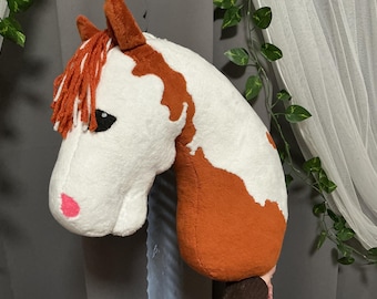 May include: A plush horse head toy featuring a white and brown spotted coat, a bright pink nose, and a textured orange yarn mane. The soft fabric construction includes pointed ears and dark eyes, creating a detailed representation of a horse.