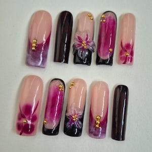 Op de afbeelding: Een set van tien kunstnagels in verschillende ontwerpen. Sommige nagels hebben een roze en paarse gradiënt, terwijl andere een 3D-bloemdesign met gouden accenten hebben. Twee nagels zijn effen donkerbruin.