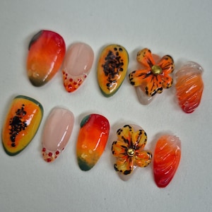 Op de afbeelding: Een verzameling van tien kunstnagels met verschillende ontwerpen. Sommige nagels hebben een verloop van oranje en rood, terwijl andere papajastukjes laten zien. Twee nagels zijn versierd met oranje bloemmotieven en één nagel heeft een wervelontwerp.