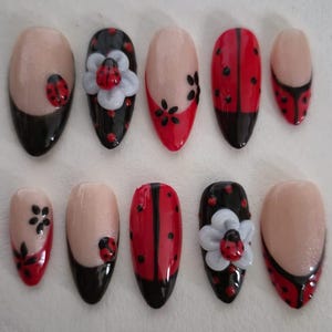 Op de afbeelding: Een set van tien kunstnagels met diverse ontwerpen, waaronder rood-zwarte lieveheersbeestjespatronen, witte 3D-bloemaccenten, zwarte bloemmotieven en Franse nageltips in rood en zwart op een nude basis met een glanzende afwerking.