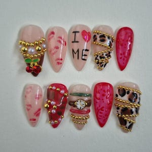 Op de afbeelding: Een set van tien amandelvormige kunstnagels met diverse ontwerpen. De ontwerpen omvatten lipafdrukken, de woorden "I ME" met een hart, luipaardprint, kersen en horlogedetails. Versieringen zijn gouden kralen en parels.