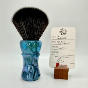 Puede incluir: Una brocha de afeitar con mango de resina azul y verde y cabeza de cerdas negras. La brocha tiene una etiqueta que dice "Soft Serve" y "Resin". La etiqueta está sujeta por un pequeño bloque de madera.