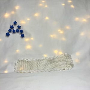 May include: A cream-colored, crocheted headband with a scalloped edge. The headband is rectangular and lies on a white surface. Blue star-shaped origami are arranged in the shape of the letter "A".