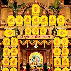 May include: A large, ornate decorative archway featuring numerous yellow, shield-shaped panels with intricate red patterns. The structure is adorned with clay pots, green foliage, and a central banner with Bengali text, framing a shrine with figures.