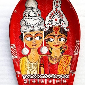 May include: A hand-painted, red woven tray featuring a couple in traditional Indian wedding attire. The artwork depicts a man and woman with detailed headwear, jewelry, and clothing. The tray has a decorative border.