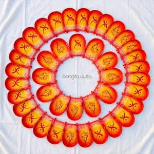 May include: A collection of small, oval-shaped clay oil lamps arranged in two concentric circles. Each lamp features a gradient of yellow and red paint, with hand-painted black symbols or Ganesha motifs. The text Sangita dutta is centered.