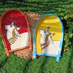 Op de afbeelding: Twee handgeschilderde bamboe trays. Eén heeft een rode achtergrond, de andere geel. Elk toont een witte koe met gouden accenten. De trays liggen op een geweven mat, tegen een achtergrond van groen gras en planten.