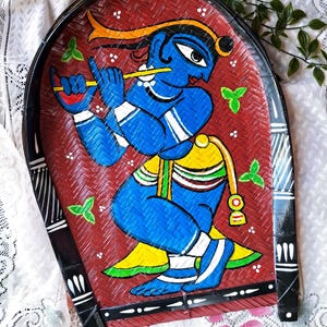 May include: Hand-painted, arched serving tray featuring a blue figure playing a flute. The figure is set against a red woven background with green leaf accents. The tray has a black border with white accents.