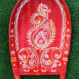 May include: A red woven tray with a decorative paisley design in white and gold. The tray has a curved shape with two small legs and is set against a green background.