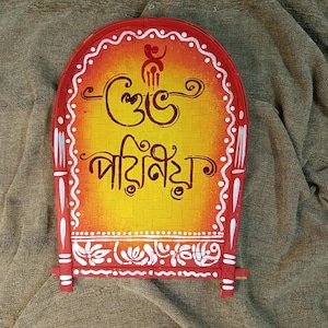 May include: A hand-painted, arched wooden plaque with Bengali script. The plaque features a yellow and orange background with white and red decorative borders. The text is in dark brown. The plaque is supported by two small wooden legs.