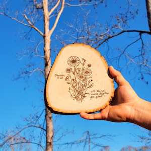 May include: A light-colored wooden slice featuring a dark, burned-in line art illustration of a wildflower bouquet. The text "Romans 8:28" is on the left, and "all things work together for good" is written on the right side of the flowers.