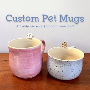 May include: Two handmade ceramic mugs with dog figurines on the rims. One mug is pink, the other is speckled blue. The mugs have the text "Custom Pet Mugs" and "A handmade mug to honor your pet!"