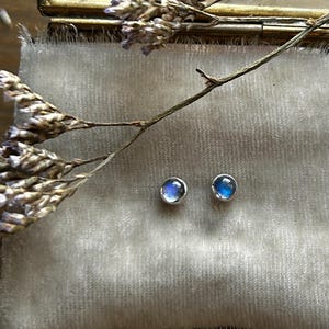 May include: A pair of small, round stud earrings featuring iridescent blue moonstones set in polished silver bezels. The earrings are displayed on a soft, light-colored velvet cushion inside a small, rectangular brass-framed glass box.