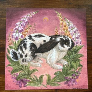 May include: A square art print on wood featuring a black and white spotted rabbit centered amidst a wreath of purple and yellow foxglove flowers. The background is a deep magenta hue with a small golden crescent moon depicted above the rabbit.