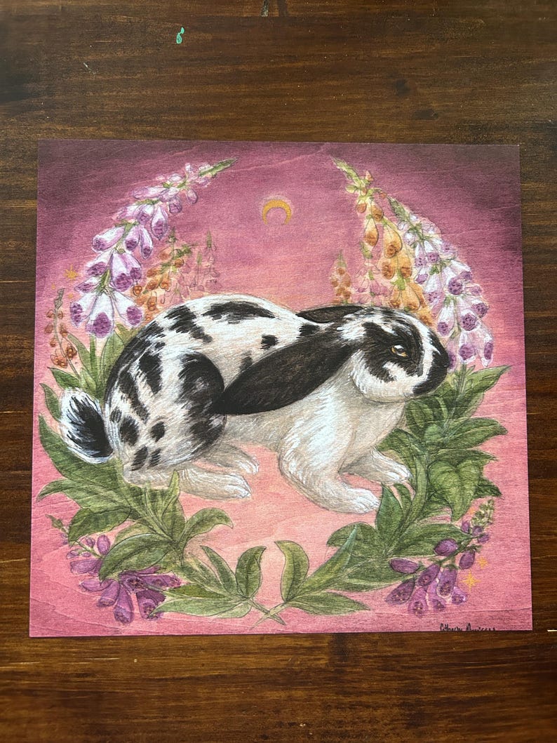 May include: A square art print on wood featuring a black and white spotted rabbit centered amidst a wreath of purple and yellow foxglove flowers. The background is a deep magenta hue with a small golden crescent moon depicted above the rabbit.