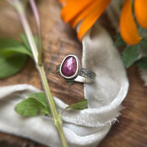 May include: A silver ring featuring a faceted, pear-shaped dark pink gemstone set in a smooth bezel. The band has a textured, etched surface with a dark oxidized finish, contrasting with the polished silver setting surrounding the central stone.