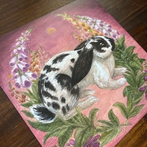 May include: A square art print featuring a black and white spotted lop-eared rabbit centered within a wreath of green leaves and purple foxglove flowers against a dusty pink background with a small yellow crescent moon in the upper section.
