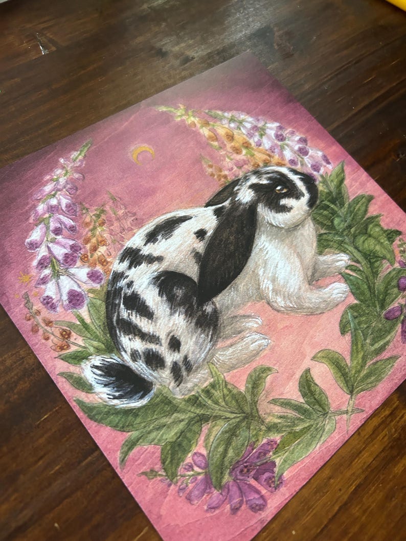 May include: A square art print featuring a black and white spotted lop-eared rabbit centered within a wreath of green leaves and purple foxglove flowers against a dusty pink background with a small yellow crescent moon in the upper section.