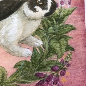 May include: A painting of a black and white rabbit with amber eyes positioned next to green leaves and purple foxglove flowers on a textured mauve background. The artist signature Catherine Morrissey is visible in the bottom right corner.