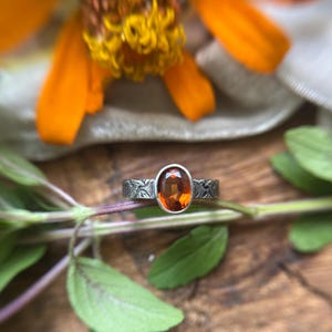 May include: A ring featuring an oval-cut orange gemstone set in a silver band with a textured, dark, etched surface pattern. The stone is faceted and clear, reflecting light, while the metal band displays a matte, oxidized finish with intricate lines.
